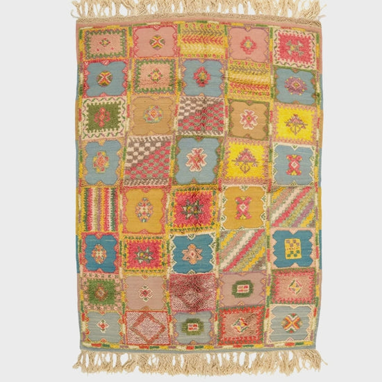 video of a rectangular patchwork-style moroccan rug with fringed top and bottom edges, featuring a multicolor grid of geometric and floral motifs in pink, yellow, blue, green, and beige tones on a white background.
