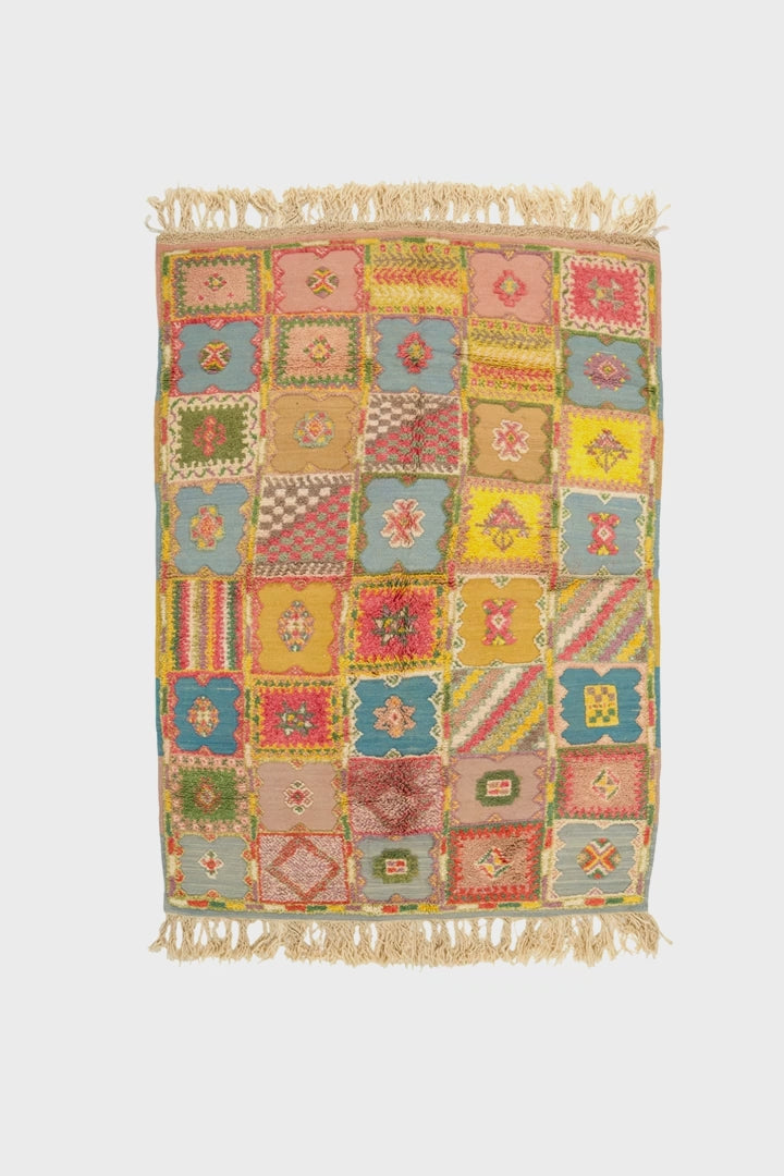 video of a rectangular patchwork-style moroccan rug with fringed top and bottom edges, featuring a multicolor grid of geometric and floral motifs in pink, yellow, blue, green, and beige tones on a white background.