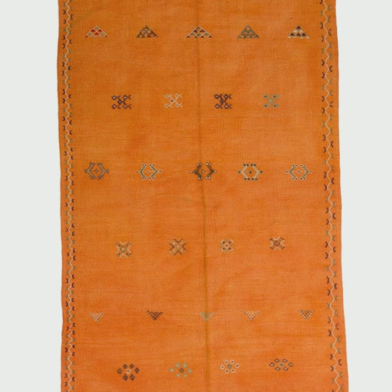video of an orange Moroccan wool rug with a geometric hand embroidered tribal motifs and white fringe, shown flat against a white background.