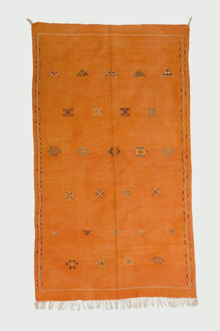 video of an orange Moroccan wool rug with a geometric hand embroidered tribal motifs and white fringe, shown flat against a white background.