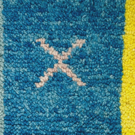 Close-up video of a textured wool moroccan rug with vertical color blocks: light blue, medium blue, yellow, and black, with small geometric motifs including a pale pink X in the center blue section.