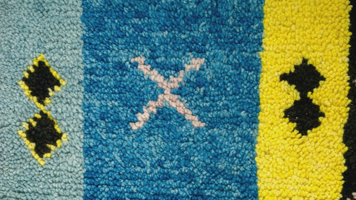 Close-up video of a textured wool moroccan rug with vertical color blocks: light blue, medium blue, yellow, and black, with small geometric motifs including a pale pink X in the center blue section.