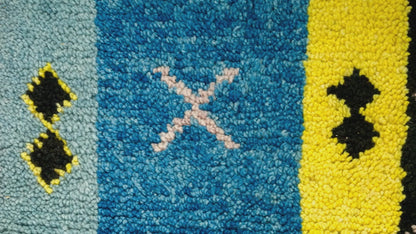 Close-up video of a textured wool moroccan rug with vertical color blocks: light blue, medium blue, yellow, and black, with small geometric motifs including a pale pink X in the center blue section.